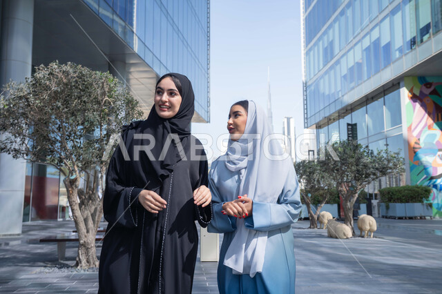 قضاء اوقات ترفيهية مع الاصدقاء ، الفخر والاعتزاز بالتقدم المعماري ، سيدتان اماراتيان تنظران النمو والتطور العمراني ، التجول واستنشاق الهواء النقي في الخارج, صديقتان عربيتان خليجيتان سعوديتان ترتديان العباءات والحجاب تنظران الى شئ ما بتعابير السرور في وقت النهار.