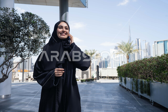 قضاء أوقات ممتعة ، سيدة اماراتية تنظر بتعابير السرور تقوم باستخدام الهاتف المحمول ، التقاط صور السيلفي عبر الجوال ، التجول في المناطق العامة, التواصل مع الأهل والأصدقاء، اجراء المكالمات الهاتفية مع العائلة، توثيق اللحظات السعيدة، امرأة عربية خليجية سعودية ترتدي العباءة والحجاب، نزهة  فى الهواء الطلق .