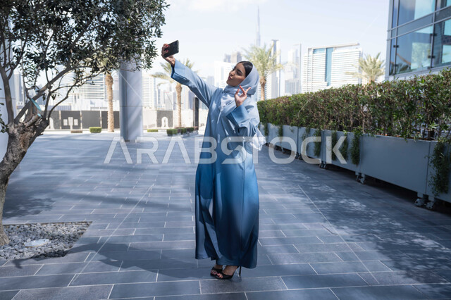 سيدة اماراتية تنظر بتعابير السرور تقوم باستخدام الهاتف المحمول ، التقاط صور السيلفي عبر الجوال ، التجول في المناطق العامة, التواصل مع الأهل والأصدقاء، اجراء مكالمات الفيديو مع العائلة، توثيق اللحظات السعيدة، امرأة عربية خليجية سعودية ترتدي العباءة والحجاب، نزهة  فى الهواء الطلق .