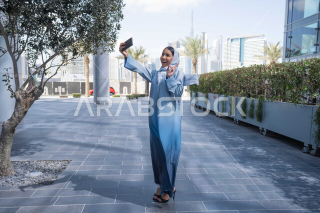 التقاط صور السيلفي عبر الجوال ، التجول في المناطق العامة, التواصل مع الأهل والأصدقاء، اجراء مكالمات الفيديو مع العائلة، توثيق اللحظات السعيدة، امرأة عربية خليجية سعودية ترتدي العباءة والحجاب، سيدة اماراتية تنظر بتعابير السرور تقوم باستخدام الهاتف المحمول ، نزهة  فى الهواء الطلق .