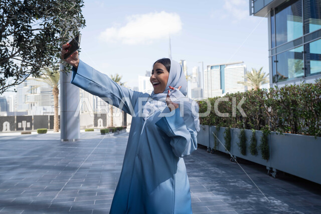 استخدام الهاتف المحمول والتقنيات الحديثه ، التجول في المناطق العامة, التواصل مع الأهل والأصدقاء، اجراء مكالمات الفيديو مع العائلة، توثيق اللحظات السعيدة، امرأة عربية خليجية سعودية ترتدي العباءة والحجاب، سيدة اماراتية تنظر بتعابير السرور تقوم باستخدام الهاتف المحمول ، نزهة  فى الهواء الطلق .