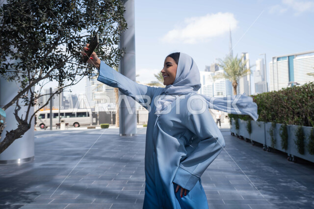 Making video calls with family, documenting happy moments, using a mobile phone and modern technology, a Saudi Arabian Gulf woman wearing an abaya and hijab, an Emirati woman looking happy using a mobile phone, a walk and a walk in the fresh air.