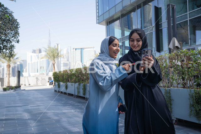 الاشارة نحو الهاتف باصبع السبابة ، الإستمتاع مع الأصدقاء,  التجول في إحدى المناطق العامة ، صديقتان عربيتان خليجيتان سعوديتان مبتسمتان تسيران معاً في نزهة ممتعة لإحدى الأماكن الترفيهية ، سيدتان اماراتيان تقومان بالتنزه في وقت العطلة ، صداقات و تعارف ، علاقات اجتماعية ، قضاء أوقات ممتعة .
