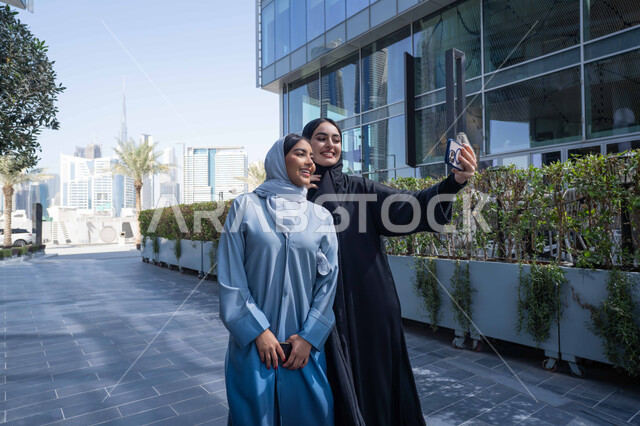 Selfies with her friend with happy gestures, making a video call, going out and getting some fresh air, documenting happy moments, fun activities and events, and spending quality time with friends. Two Saudi Arabian Gulf friends wearing abayas and hijab. Two Emirati women looking with happy expressions, one of them using her mobile phone.