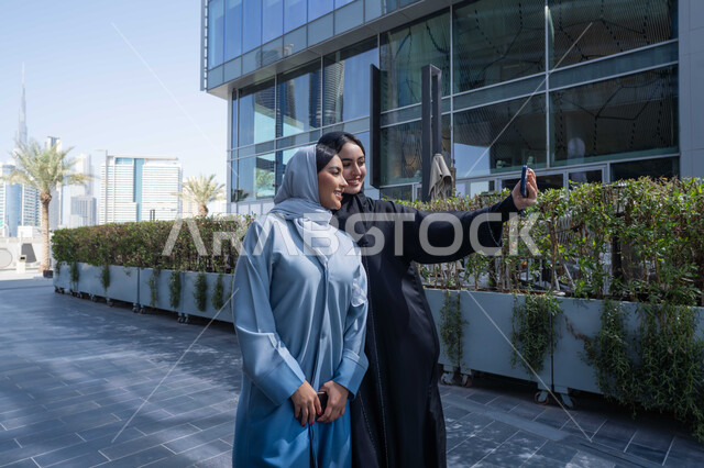 استخدام الجوال والتقنيات الحديثه، اجراء مكالمة فيديو، الخروج واستنشاق الهواء النقي ، توثيق اللحظات السعيدة، فعاليات وأنشطة ترفيهية وقضاء وقت ممتع مع الاصدقاء ، صديقتان عربيتان خليجيتان سعوديتان ترتديان العباءات والحجاب، سيدتان اماراتيان تنظران بتعابير السرور ، احداهما تقوم باستخدام الهاتف المحمول .