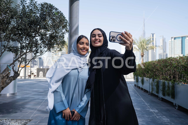 الخروج واستنشاق الهواء النقي ، توثيق اللحظات السعيدة، استخدام الهاتف المحمول والتقنيات الحديثه، اجراء مكالمة فيديو ، فعاليات وأنشطة ترفيهية وقضاء وقت ممتع مع الاصدقاء ، صديقتان عربيتان خليجيتان سعوديتان ترتديان العباءات والحجاب، سيدتان اماراتيان تنظران بتعابير السرور ، احداهما تقوم باستخدام الهاتف المحمول .