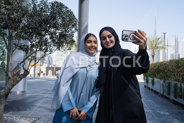 توثيق اللحظات السعيدة، استخدام الهاتف المحمول والتقنيات الحديثه، اجراء مكالمة فيديو ، فعاليات وأنشطة ترفيهية وقضاء وقت ممتع مع الاصدقاء ، صديقتان عربيتان خليجيتان سعوديتان ترتديان العباءات والحجاب، سيدتان اماراتيان تنظران بتعابير السرور ، احداهما تقوم باستخدام الهاتف المحمول .