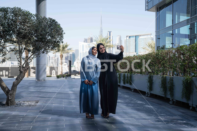 Going out in the fresh air during the day, taking souvenir photos, making a video call, having fun and having a good time with friends, two veiled Saudi Arabian Gulf Arab friends wearing abayas and hijab, two Emirati women looking with happy expressions, one of them using her mobile phone.