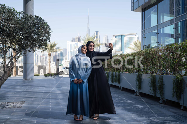 التقاط  صور تذكارية ، اجراء مكالمة فيديو ، فعاليات وأنشطة ترفيهية وقضاء وقت ممتع مع الاصدقاء ، الخروج في الهواء الطلق في وقت النهار, صديقتان عربيتان خليجيتان سعوديتان ترتديان العباءات والحجاب، سيدتان اماراتيان تنظران بتعابير السرور ، احداهما تقوم باستخدام الهاتف المحمول .