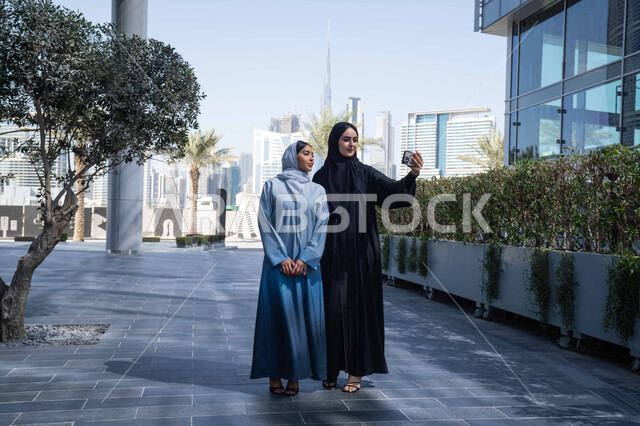 توثيق اللحظات السعيدة، اجراء مكالمة فيديو ، فعاليات وأنشطة ترفيهية وقضاء وقت ممتع مع الاصدقاء ، الخروج في الهواء الطلق في وقت النهار, صديقتان عربيتان خليجيتان سعوديتان ترتديان العباءات والحجاب، سيدتان اماراتيان تنظران بتعابير السرور ، احداهما تقوم باستخدام الهاتف المحمول .