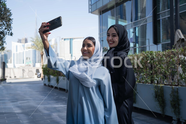 Enjoying taking selfies with your mobile phone, going out in the fresh air during the day, two veiled Saudi Arabian Gulf friends wearing abayas and hijabs, two Emirati women looking happy, one of them using her mobile phone, recreational activities and events, and spending a fun time with friends.