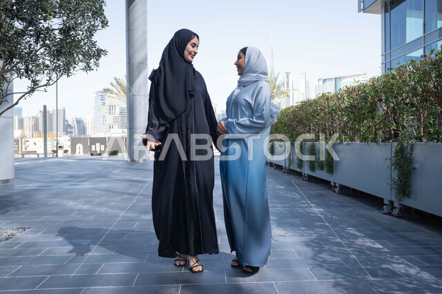 المشي فى الخارج  ، مفهوم الأناقة والاهتمام بالمظهر الخارجي ، الوقوف بثقة في شوارع المدينة، الاستمتاع والتجول واستنشاق الهواء النقي في وقت النهار، صديقتان عربيتان خليجيتان سعوديتان محجبتان ترتديان العباءات والحجاب ، زميلتان اماراتيان تنظران الى بعضهما بتعابير السرور.