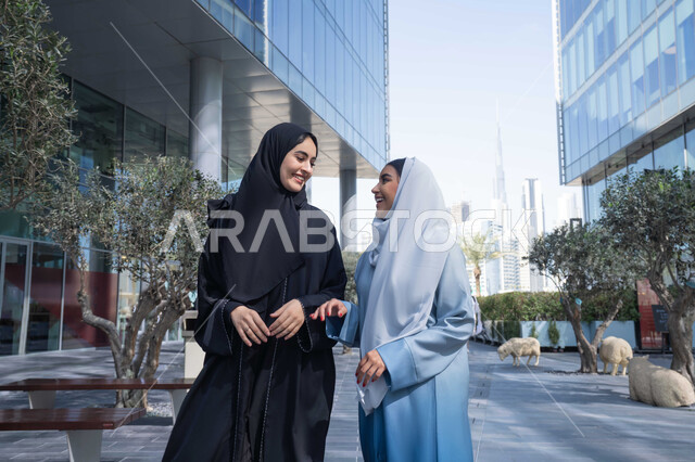 الوقوف بثقة في شوارع المدينة، الاستمتاع والتجول واستنشاق الهواء النقي في وقت النهار، صديقتان عربيتان خليجيتان سعوديتان ترتديان العباءات والحجاب ،زميلتان اماراتيان تنظران الى بعضهما بتعابير السرور، مفهوم الأناقة والاهتمام بالمظهر الخارجي .
