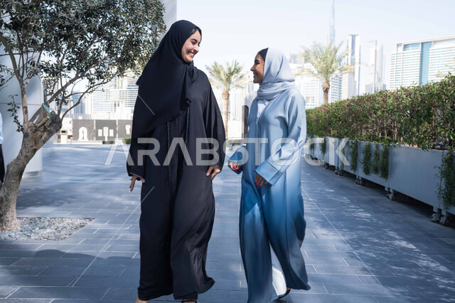 Enjoying, walking, and breathing fresh air during the day. Two Saudi Arabian Gulf friends wearing abayas and hijabs, and two Emirati colleagues looking at each other with happy expressions. Standing confidently on city streets. The concept of elegance and attention to one's appearance.