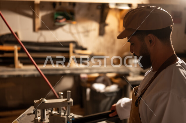 أدوات ومعدات صناعية ثقيلة لقص الخشب، مفهوم الموهبة و الإبداع، ورشة عمل فنية، رجل عربي خليجي عماني مبتسم يرتدي الدشداشة والعمامة مع المريلة يقوم بصناعة اطار للوحات، الشغف في ممارسة الهوايات المفضلة والفنون اليدوية