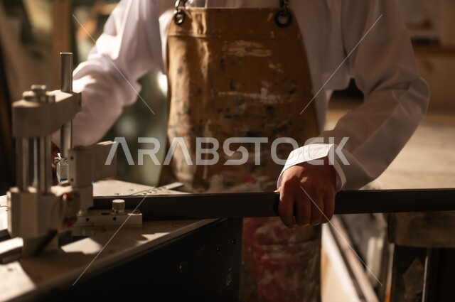 أدوات ومعدات صناعية ثقيلة لقص الخشب، مفهوم الموهبة و الإبداع، ورشة عمل فنية، رجل عربي خليجي عماني مبتسم يرتدي الدشداشة والعمامة مع المريلة يقوم بصناعة اطار للوحات، الشغف في ممارسة الهوايات المفضلة والفنون اليدوية