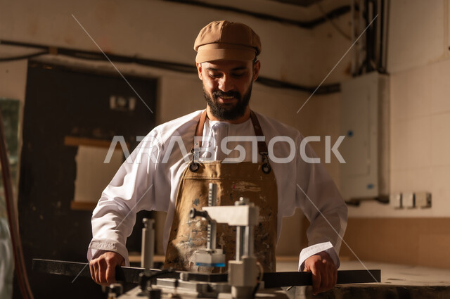 أدوات ومعدات صناعية ثقيلة لقص الخشب، مفهوم الموهبة و الإبداع، ورشة عمل فنية، رجل عربي خليجي عماني مبتسم يرتدي الدشداشة والعمامة مع المريلة يقوم بصناعة اطار للوحات، الشغف في ممارسة الهوايات المفضلة والفنون اليدوية