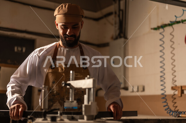 معدات وأدوات صناعية ثقيلة لقص الخشب، مفهوم الموهبة و الإبداع، ورشة عمل فنية، رجل عربي خليجي عماني مبتسم يرتدي الدشداشة والعمامة مع المريلة يقوم بصناعة اطار للوحات، الشغف في ممارسة الهوايات المفضلة والفنون اليدوية