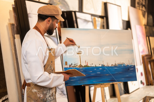 استخدام الريشة للرسم على اللوحة، مفهوم الموهبة و الإبداع، ورشة عمل فنية، رسام عربي خليجي عماني يرتدي الدشداشة والعمامة مع مريلة الرسم يقوم برسم الطبيعة العمانية، الشغف في ممارسة الهوايات المفضلة والفنون اليدوية، تعابير وايماءات الاندماج والسرور