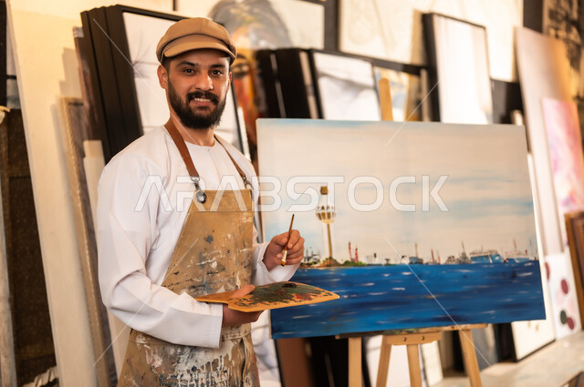 استخدام الريشة للرسم على اللوحة، مفهوم الموهبة و الإبداع، ورشة عمل فنية، رسام عربي خليجي عماني يرتدي الدشداشة والعمامة مع مريلة الرسم يقوم برسم الطبيعة العمانية، الشغف في ممارسة الهوايات المفضلة والفنون اليدوية، تعابير وايماءات الاندماج والسرور