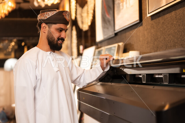 طباعة اللوحات والرسومات باستخدام الطابعة، مفهوم الموهبة و الإبداع، ورشة عمل فنية، رجل عربي خليجي عماني مبتسم يرتدي الدشداشة والعمامة يقف بايماءات السرور، الشغف في ممارسة الهوايات المفضلة والفنون اليدوية