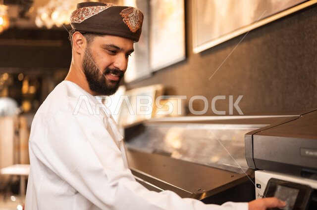 Printing paintings and drawings using a printer, the concept of talent and creativity, an art workshop, a smiling Arab Gulf Omani man wearing a dishdasha and turban standing with gestures of pleasure, passion for practicing favorite hobbies and handicrafts