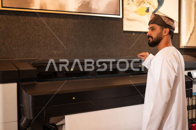 طباعة اللوحات والرسومات باستخدام الطابعة، مفهوم الموهبة و الإبداع، ورشة عمل فنية، رجل عربي خليجي عماني مبتسم يرتدي الدشداشة والعمامة يقف بايماءات السرور، الشغف في ممارسة الهوايات المفضلة والفنون اليدوية