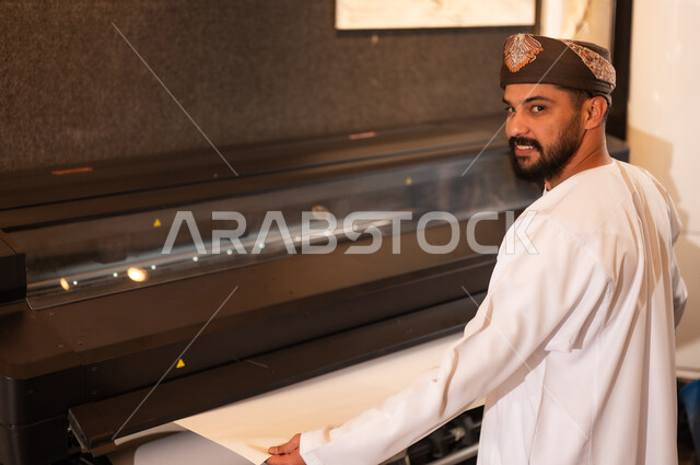 طباعة اللوحات والرسومات باستخدام الطابعة، مفهوم الموهبة و الإبداع، ورشة عمل فنية، رجل عربي خليجي عماني مبتسم يرتدي الدشداشة والعمامة يقف بايماءات السرور، الشغف في ممارسة الهوايات المفضلة والفنون اليدوية
