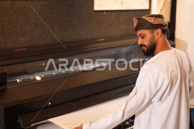 طباعة اللوحات والرسومات باستخدام الطابعة، مفهوم الموهبة و الإبداع، ورشة عمل فنية، رجل عربي خليجي عماني مبتسم يرتدي الدشداشة والعمامة يقف بايماءات السرور، الشغف في ممارسة الهوايات المفضلة والفنون اليدوية