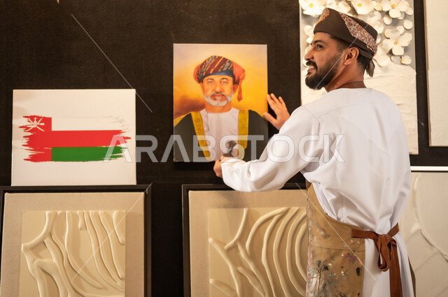 A drawing of Sultan Haitham bin Tariq, an art workshop, an Arab Gulf Omani artist wearing a dishdasha, turban and apron hanging a plaque of His Majesty the King, passion for practicing favorite hobbies and handicrafts, expressions and gestures of integration and pleasure, the concept of talent and creativity