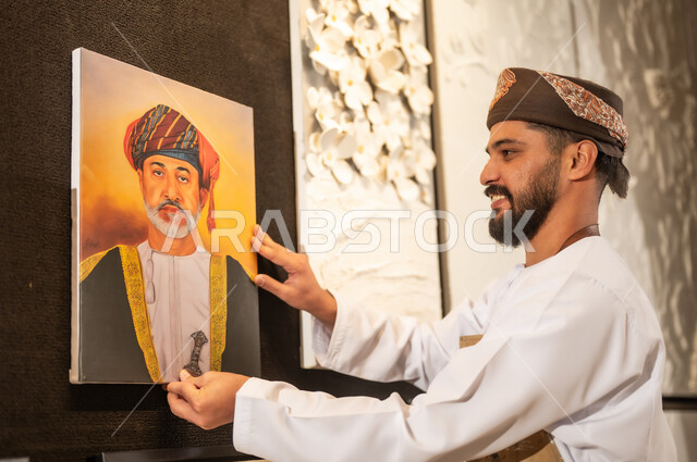 الشغف في ممارسة الهوايات المفضلة والفنون اليدوية، تعابير وايماءات الاندماج والسرور، مفهوم الموهبة و الإبداع، رسمة للسلطان هيثم بن طارق، ورشة عمل فنية، رسام عربي خليجي عماني يرتدي الدشداشة والعمامة ومريلة الرسم يقوم بتعليق لحة جلالة الملك المعظم