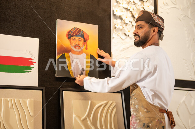 A drawing of Sultan Haitham bin Tariq, an art workshop, an Arab Gulf Omani artist wearing a dishdasha, turban and apron hanging a plaque of His Majesty the King, passion for practicing favorite hobbies and handicrafts, expressions and gestures of integration and pleasure, the concept of talent and creativity