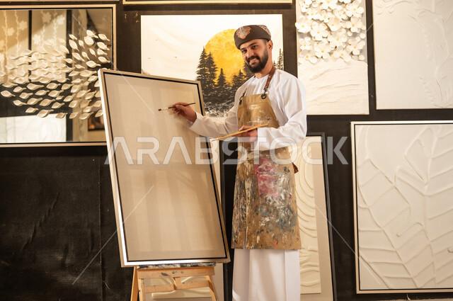 تعابير وايماءات الاندماج والسرور، استخدام الريشة للرسم على اللوحة، مفهوم الموهبة و الإبداع، ورشة عمل فنية، رسام عربي خليجي عماني يرتدي الدشداشة والعمامة مع مريلة الرسم يستعد لرسم شيء ما، الشغف في ممارسة الهوايات المفضلة والفنون اليدوية