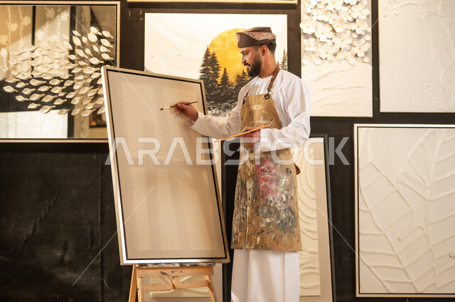 تعابير وايماءات الاندماج والسرور، استخدام الريشة للرسم على اللوحة، مفهوم الموهبة و الإبداع، ورشة عمل فنية، رسام عربي خليجي عماني يرتدي الدشداشة والعمامة مع مريلة الرسم يستعد لرسم شيء ما، الشغف في ممارسة الهوايات المفضلة والفنون اليدوية