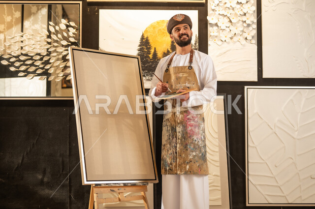 Expressions and gestures of integration and pleasure, using a brush to draw on a canvas, the concept of talent and creativity, an art workshop, an Arab Gulf Omani painter wearing a dishdasha and turban with a painting apron preparing to draw something, passion for practicing favorite hobbies and handicrafts