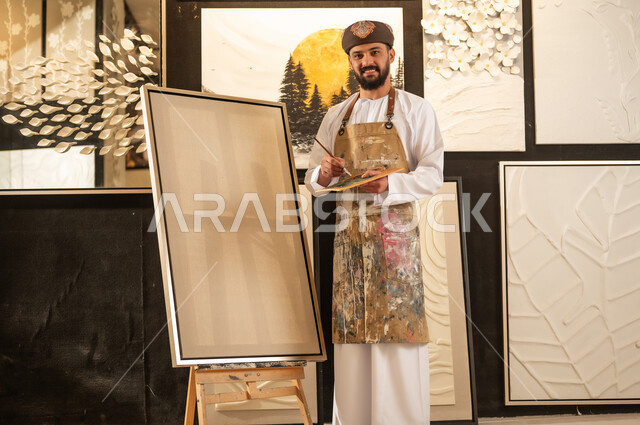 تعابير وايماءات الاندماج والسرور، استخدام الريشة للرسم على اللوحة، مفهوم الموهبة و الإبداع، ورشة عمل فنية، رسام عربي خليجي عماني يرتدي الدشداشة والعمامة مع مريلة الرسم يستعد لرسم شيء ما، الشغف في ممارسة الهوايات المفضلة والفنون اليدوية
