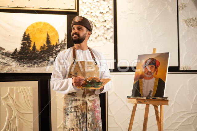 لوحة للسلطان هيثم بن طارق، ورشة عمل فنية, رسام عربي خليجي عماني يرتدي الدشداشة والعمامة ومريلة الرسم يقوم برسم جلالة الملك المعظم، الشغف في ممارسة الهوايات المفضلة والفنون اليدوية، تعابير وايماءات الاندماج والسرور، مفهوم الموهبة و الإبداع