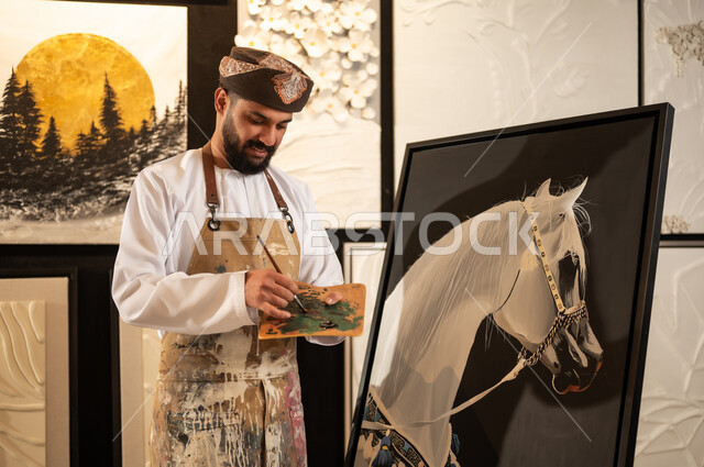 ممارسة الهوايات المفضلة والفنون اليدوية، تعابير وايماءات الشغف والاندماج والسرور، استخدام الريشة للرسم على اللوحة، مفهوم الموهبة و الإبداع، ورشة عمل فنية، رسام عربي خليجي عماني يرتدي الدشداشة والعمامة مع مريلة الرسم يقوم برسم حصان ابيض عربي اصيل