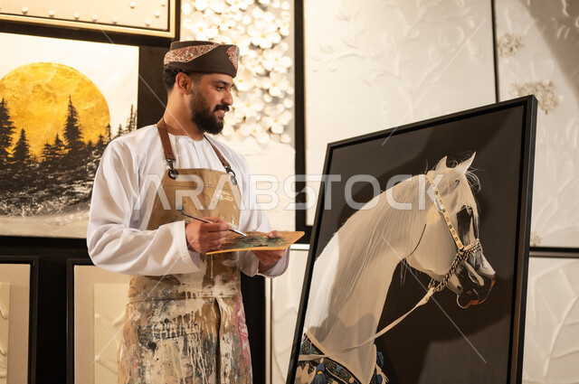 Passion for practicing favorite hobbies and handicrafts, expressions and gestures of integration and pleasure, using a brush to draw on a canvas, the concept of talent and creativity, an art workshop, an Arab Gulf Omani painter wearing a dishdasha and turban with a painting apron drawing a purebred white Arabian horse