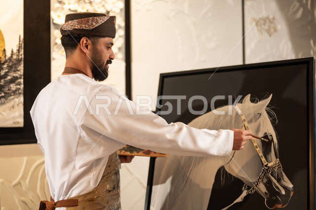 Practicing favorite hobbies and handicrafts, expressions and gestures of passion, integration and pleasure, using a brush to draw on a canvas, the concept of talent and creativity, an art workshop, an Arab Gulf Omani painter wearing a dishdasha and turban with a painting apron drawing a purebred white Arabian horse