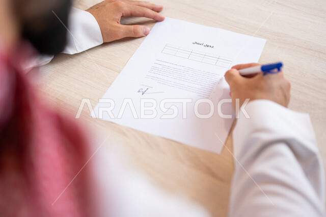 رجل عربي خليجي سعودي يسمك بيده قلم ويقوم بتجهيز جدول الأعمال، سكرتير يدير الاعمال الورقية، تنظيم جدول الاعمال والمهام، أعمال مكتبية