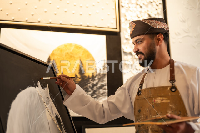 ممارسة الهوايات المفضلة والفنون اليدوية، تعابير وايماءات الشغف والاندماج والسرور، استخدام الريشة للرسم على اللوحة، مفهوم الموهبة و الإبداع، ورشة عمل فنية، رسام عربي خليجي عماني يرتدي الدشداشة والعمامة مع مريلة الرسم يقوم برسم حصان ابيض عربي اصيل