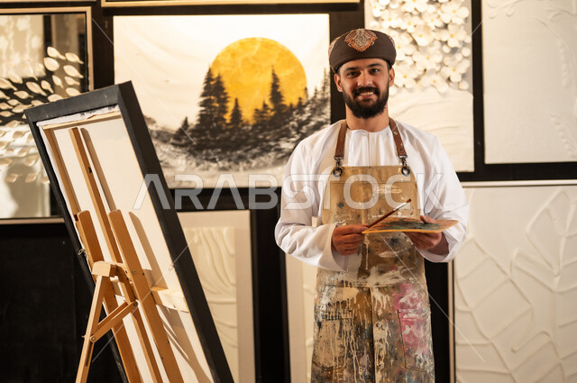 تعابير وايماءات الاندماج والسرور، استخدام الريشة للرسم على اللوحة، مفهوم الموهبة و الإبداع، ورشة عمل فنية، رسام عربي خليجي عماني يرتدي الدشداشة والعمامة مع مريلة الرسم يستعد لرسم شيء ما، الشغف في ممارسة الهوايات المفضلة والفنون اليدوية