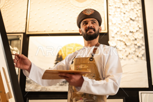 استخدام الريشة للرسم على اللوحة، مفهوم الموهبة و الإبداع، ورشة عمل فنية، رسام عربي خليجي عماني يرتدي الدشداشة والعمامة مع مريلة الرسم يستعد لرسم شيء ما، الشغف في ممارسة الهوايات المفضلة والفنون اليدوية، تعابير وايماءات الاندماج والسرور