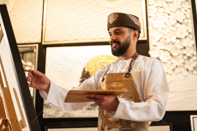 استخدام الريشة للرسم على اللوحة، مفهوم الموهبة و الإبداع، ورشة عمل فنية، رسام عربي خليجي عماني يرتدي الدشداشة والعمامة مع مريلة الرسم يستعد لرسم شيء ما، الشغف في ممارسة الهوايات المفضلة والفنون اليدوية، تعابير وايماءات الاندماج والسرور