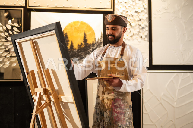 تعابير وايماءات الاندماج والسرور، استخدام الريشة للرسم على اللوحة، مفهوم الموهبة و الإبداع، ورشة عمل فنية، رسام عربي خليجي عماني يرتدي الدشداشة والعمامة مع مريلة الرسم يستعد لرسم شيء ما، الشغف في ممارسة الهوايات المفضلة والفنون اليدوية
