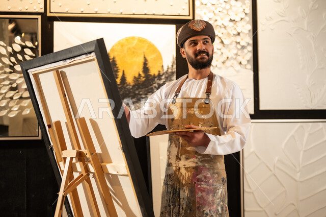 استخدام الريشة للرسم على اللوحة، مفهوم الموهبة و الإبداع، ورشة عمل فنية، رسام عربي خليجي عماني يرتدي الدشداشة والعمامة مع مريلة الرسم يستعد لرسم شيء ما، الشغف في ممارسة الهوايات المفضلة والفنون اليدوية، تعابير وايماءات الاندماج والسرور