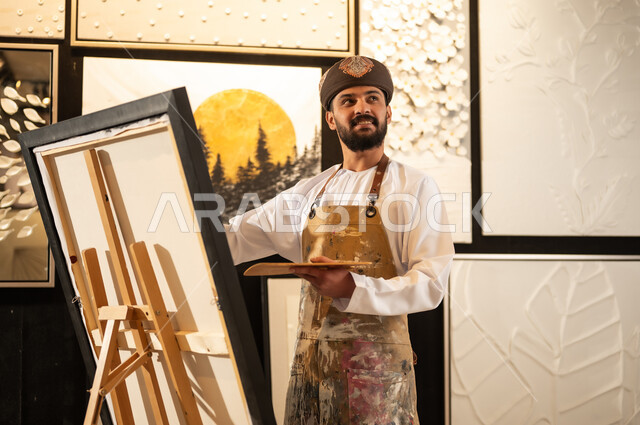 استخدام الريشة للرسم على اللوحة، مفهوم الموهبة و الإبداع، ورشة عمل فنية، رسام عربي خليجي عماني يرتدي الدشداشة والعمامة مع مريلة الرسم يستعد لرسم شيء ما، الشغف في ممارسة الهوايات المفضلة والفنون اليدوية، تعابير وايماءات الاندماج والسرور