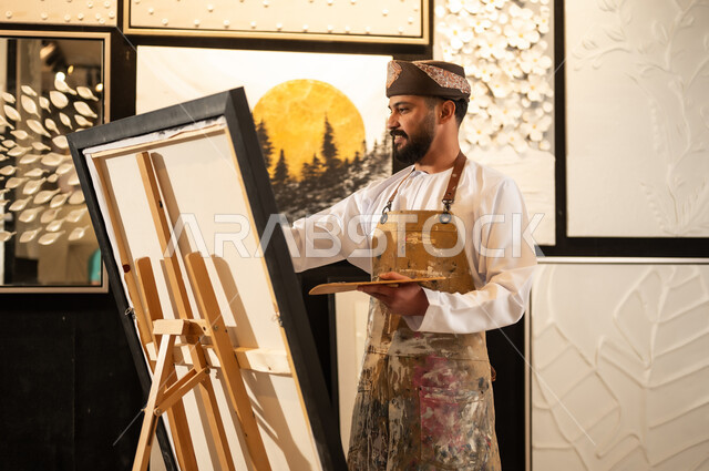 تعابير وايماءات الاندماج والسرور، استخدام الريشة للرسم على اللوحة، مفهوم الموهبة و الإبداع، ورشة عمل فنية، رسام عربي خليجي عماني يرتدي الدشداشة والعمامة مع مريلة الرسم يستعد لرسم شيء ما، الشغف في ممارسة الهوايات المفضلة والفنون اليدوية