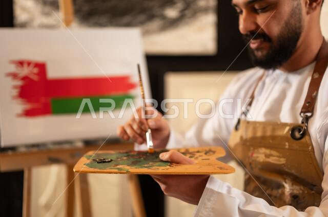 ذكرى تأسيس الدولة 1965، إحياء اليوم الوطني العماني 18 نوفمبر، ورشة عمل فنية، رسام عربي خليجي عماني يرتدي الدشداشة والعمامة مع مريلة الرسم يقوم برسم علم سلطنة عمان، ممارسة الهوايات المفضلة والفنون اليدوية، مفهوم الموهبة و الإبداع وحب الوطن والانتماء إليه